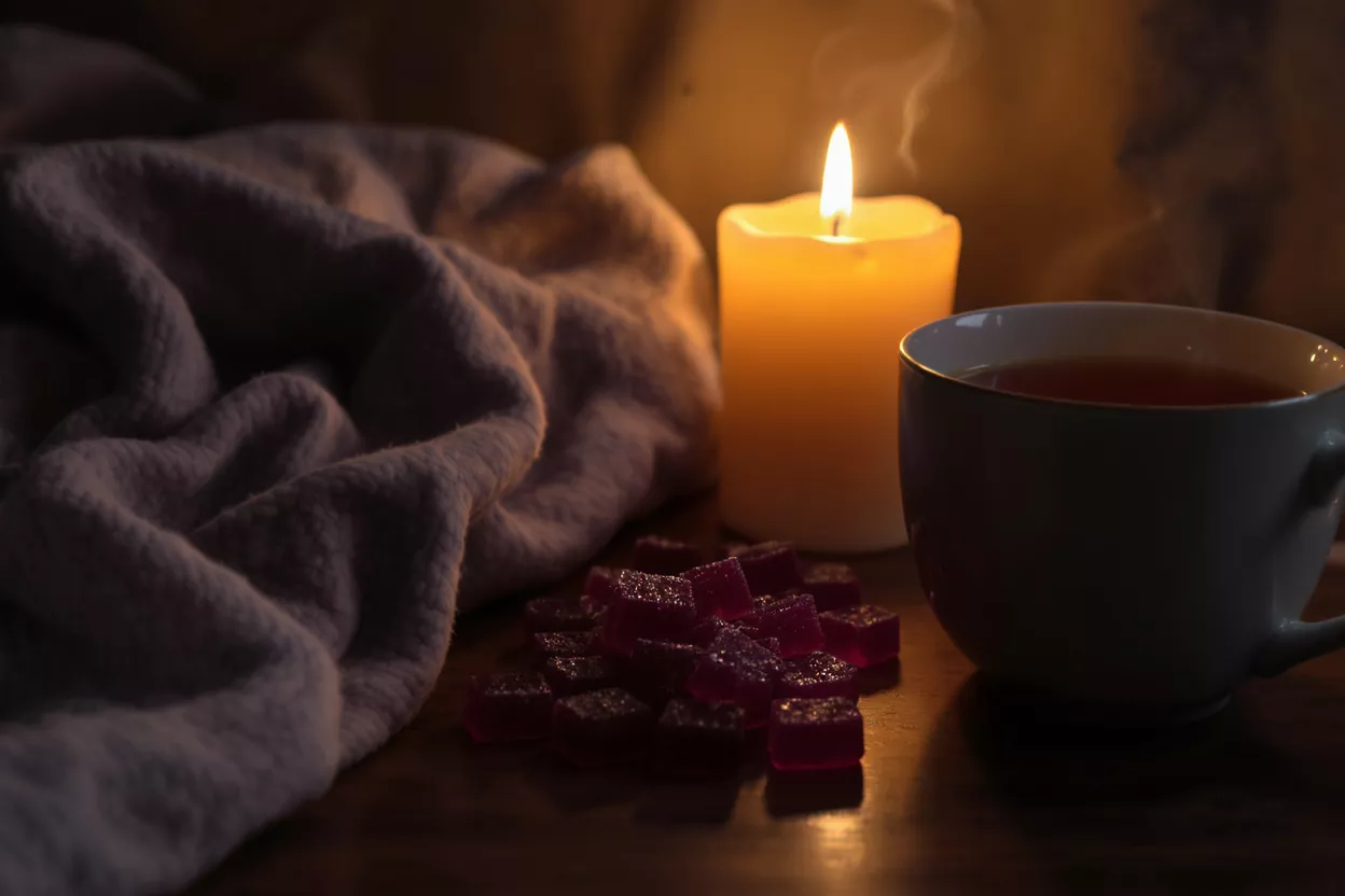 Escena cálida con una vela encendida, té humeante y gominolas moradas sobre una superficie de madera.