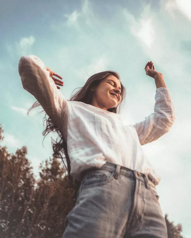 Young woman joyfully posing outdoors in sunny, natural setting with clear sky and trees.
