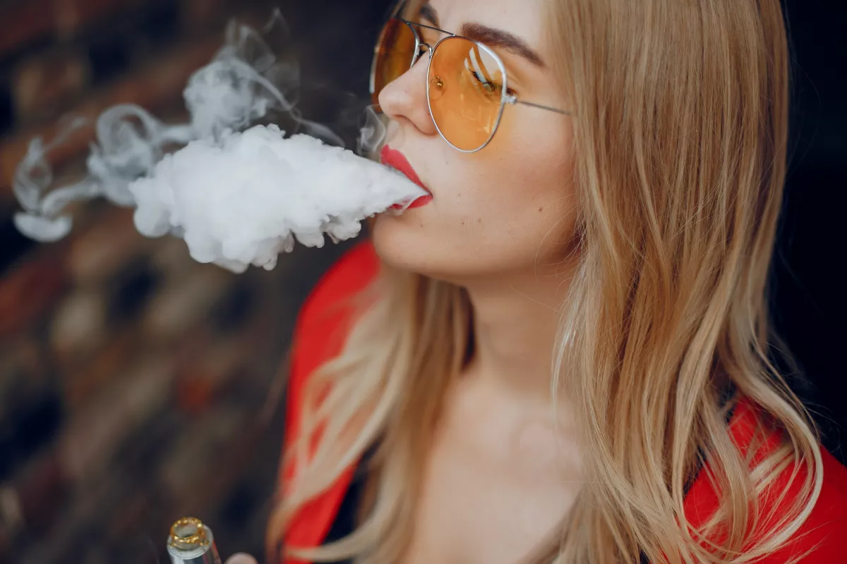 Blonde woman in sunglasses exhales vapor from e-cigarette outdoors, showcasing style and vape culture.