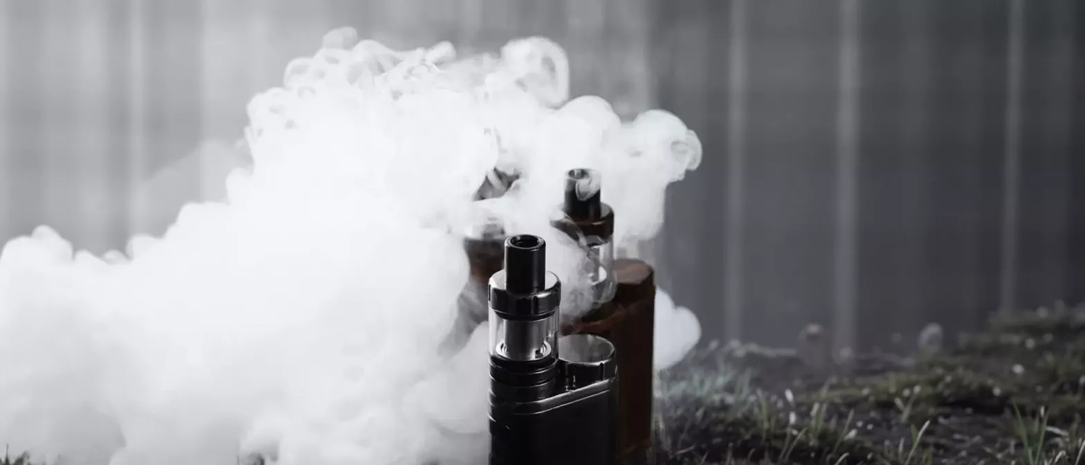 Three vape devices surrounded by thick white smoke on a dark background.