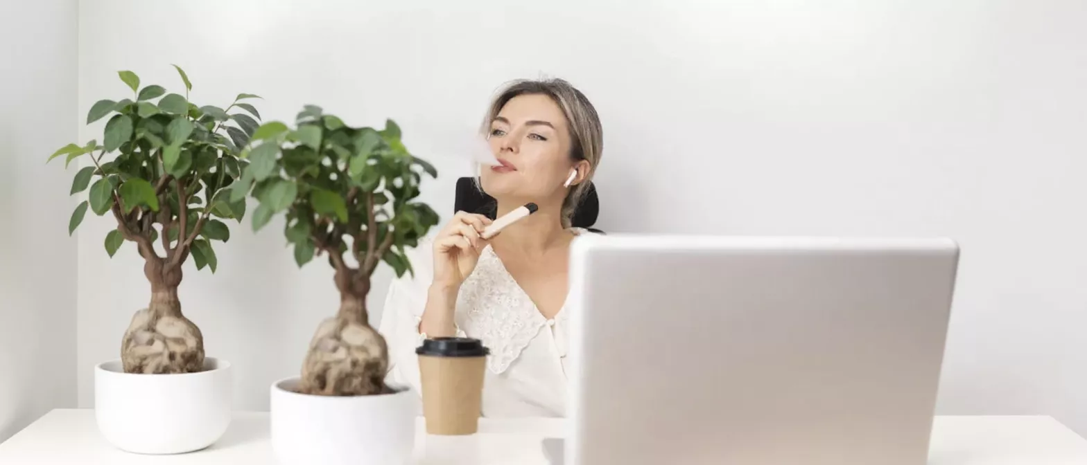 Woman vaping at a white desk with two bonsai trees and a laptop.