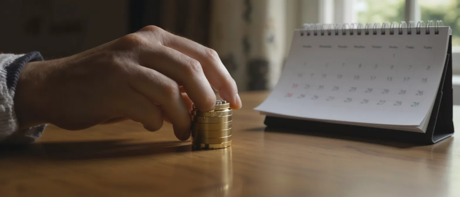 A hand stacking golden coins next to a desk calendar on a wooden table.