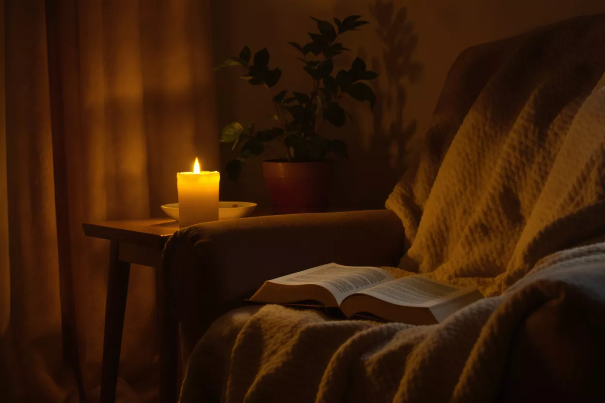 A lit candle, open book, and blanket on an armchair in a dimly lit room.