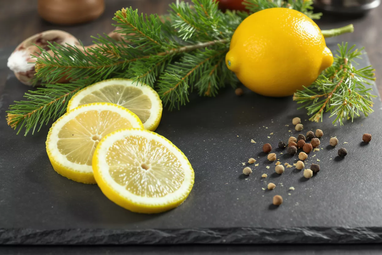 Whole lemon, sliced lemons, spruce branch, and peppercorns on a dark slate surface.