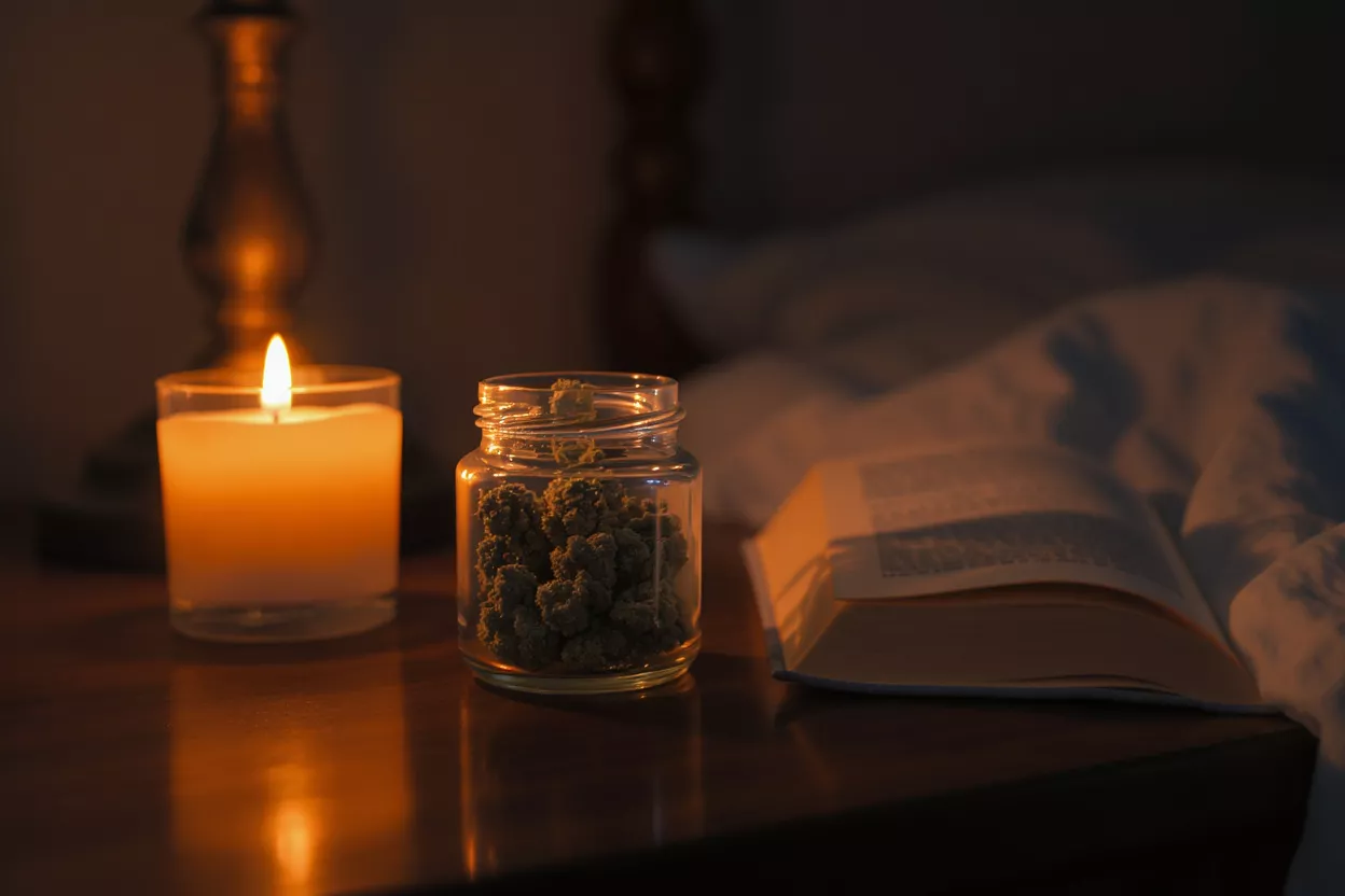 Une bougie allumée, un bocal de cannabis et un livre ouvert sur une table de chevet.