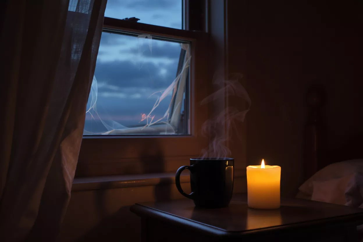 Steaming coffee mug and lit candle on a table by a window at dusk.