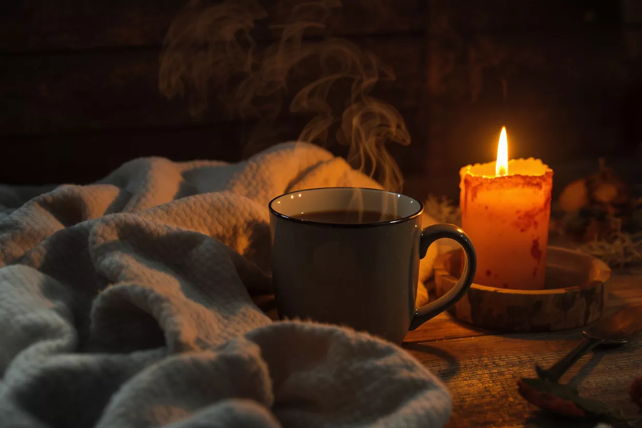 Steaming mug of coffee next to a lit candle and a warm blanket.