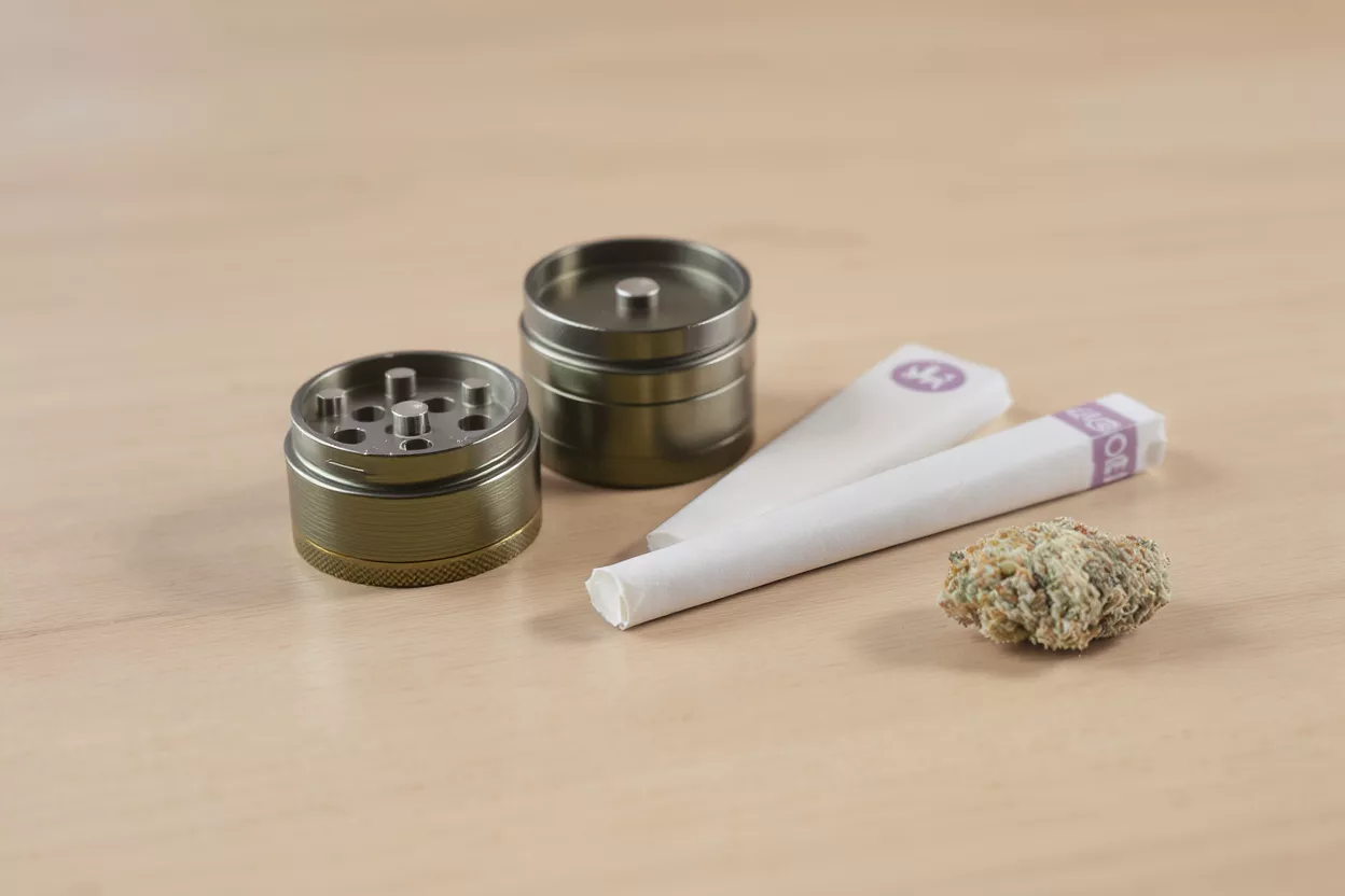 A metal grinder, two white pre-roll cones, and a cannabis bud on a wooden surface.